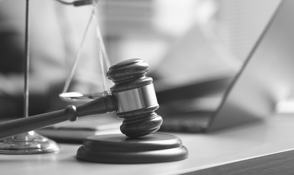Black-and-white photo of a wooden gavel on its sound block beside the scales of justice, with a laptop blurred in the background.