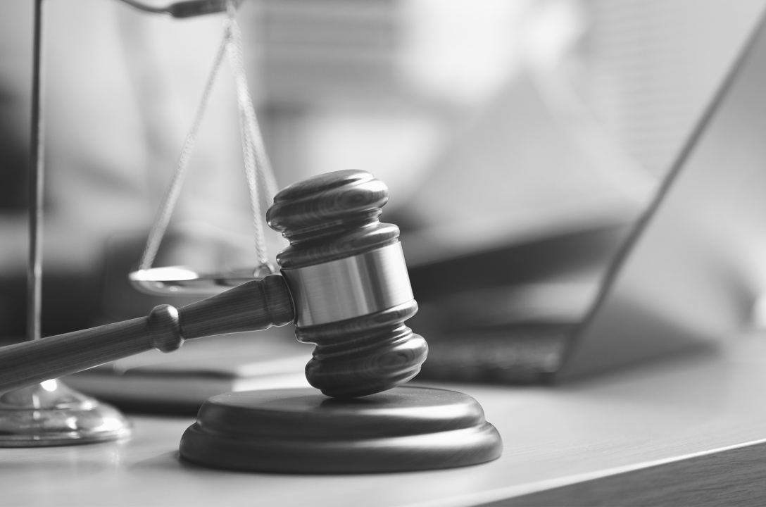 Black-and-white photo of a wooden gavel on its sound block beside the scales of justice, with a laptop blurred in the background.
